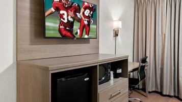 A hotel room with a flat-screen TV mounted on the wall, a desk with a chair, a microwave, and a small cabinet.