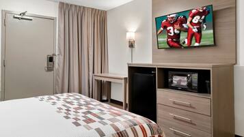 A hotel room with a bed, a desk, a refrigerator, a microwave, and a television displaying a football game.