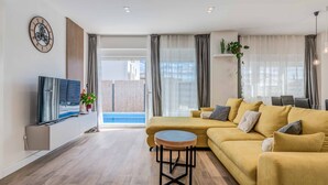 Ein modernes Wohnzimmer mit einer Couch, einem runden Couchtisch, einem Flachbildfernseher und einem großen Fenster mit Vorhängen.