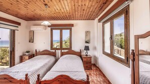 Quarto com duas camas de madeira, um ventilador de teto, mesa de cabeceira com abajur e janelas amplas com vista para o exterior.