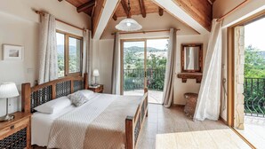 A bedroom with a wooden ceiling, a bed with a headboard, a nightstand, a lamp, a window, and a balcony with a view of greenery. - Villa Sweet Memories (Argaka)