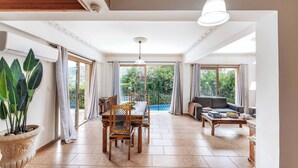A spacious living area with a dining table, chairs, a sofa, and a coffee table. There is a large potted plant, a ceiling fan, and a view of the outdoors through large windows. - Villa Sweet Memories (Argaka)