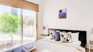 A hotel room with a large bed, a view of a garden, and a painting on the wall. - Sea Breeze Hideaway - Three Bedroom Villa, Sleeps 6 (Latchi)