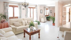 A spacious living room with a beige sofa, a wooden coffee table, and a chandelier. There are two armchairs and a potted plant. - Villa Nerja Angeles - Three Bedroom Villa, Sleeps 6 (Nerja)