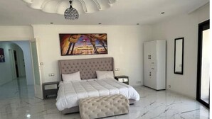 Un dormitorio moderno con una cama grande, un reposapiés tapizado, un cuadro sobre la cama y un armario blanco.