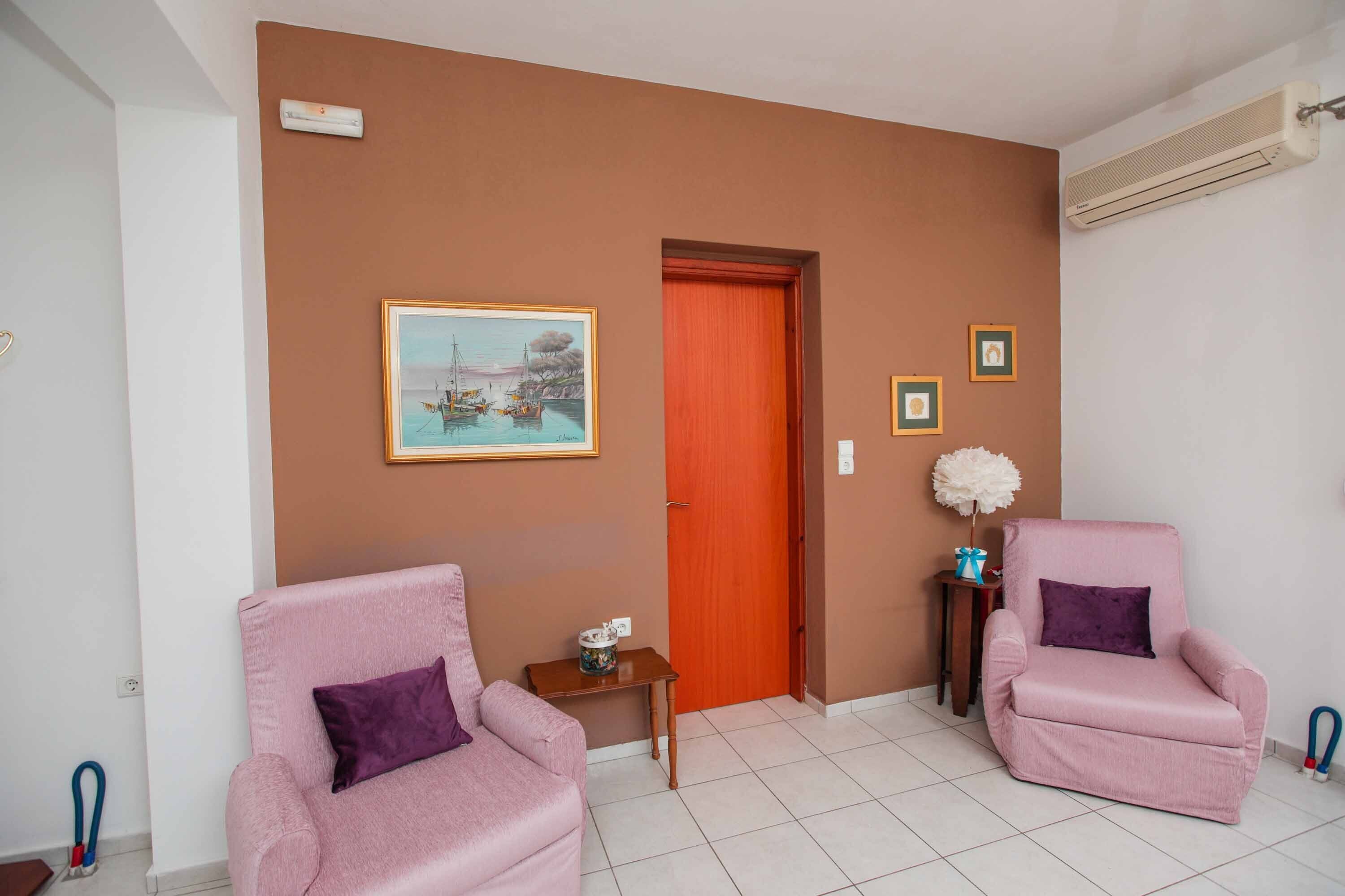 Um quarto com duas poltronas cor-de-rosa, uma porta de madeira, um quadro emoldurado e uma pequena mesa com um vaso.