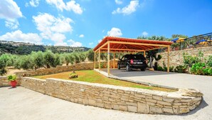 A covered parking area with a stone wall, a car, and a view of olive trees and hills.