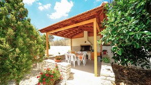 A covered outdoor dining area with a stone wall, white chairs, and a grill.
