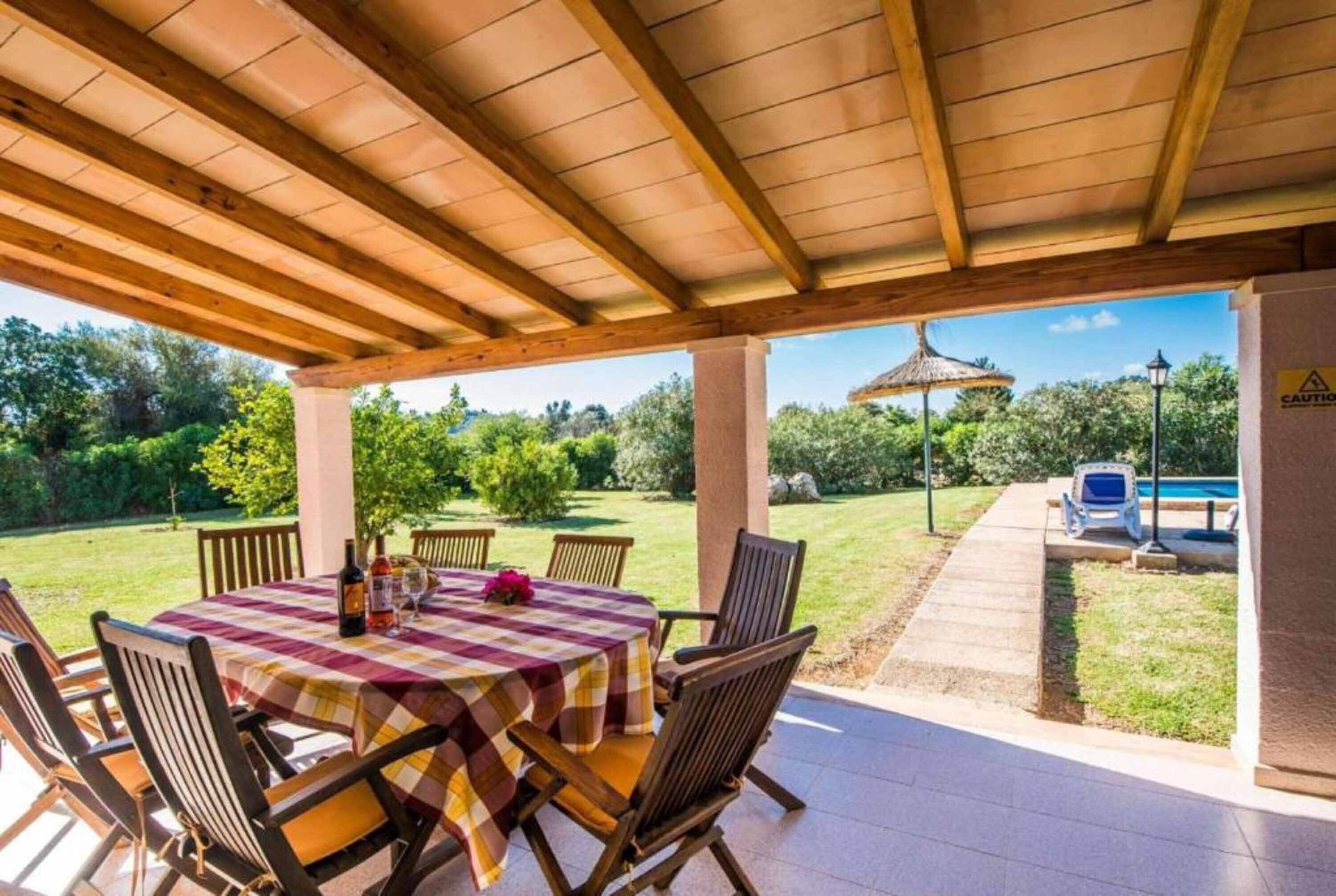 Terrassa exterior amb taula preparada per a dinar, envoltada de cadires i una zona coberta amb vista a una piscina i vegetació.