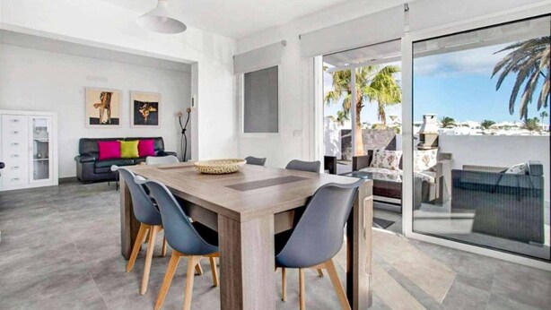 Un salon moderne avec une grande table à manger en bois, des chaises grises, un canapé noir agrémenté de coussins colorés et une porte en verre donnant sur un balcon avec vue sur un palmier.