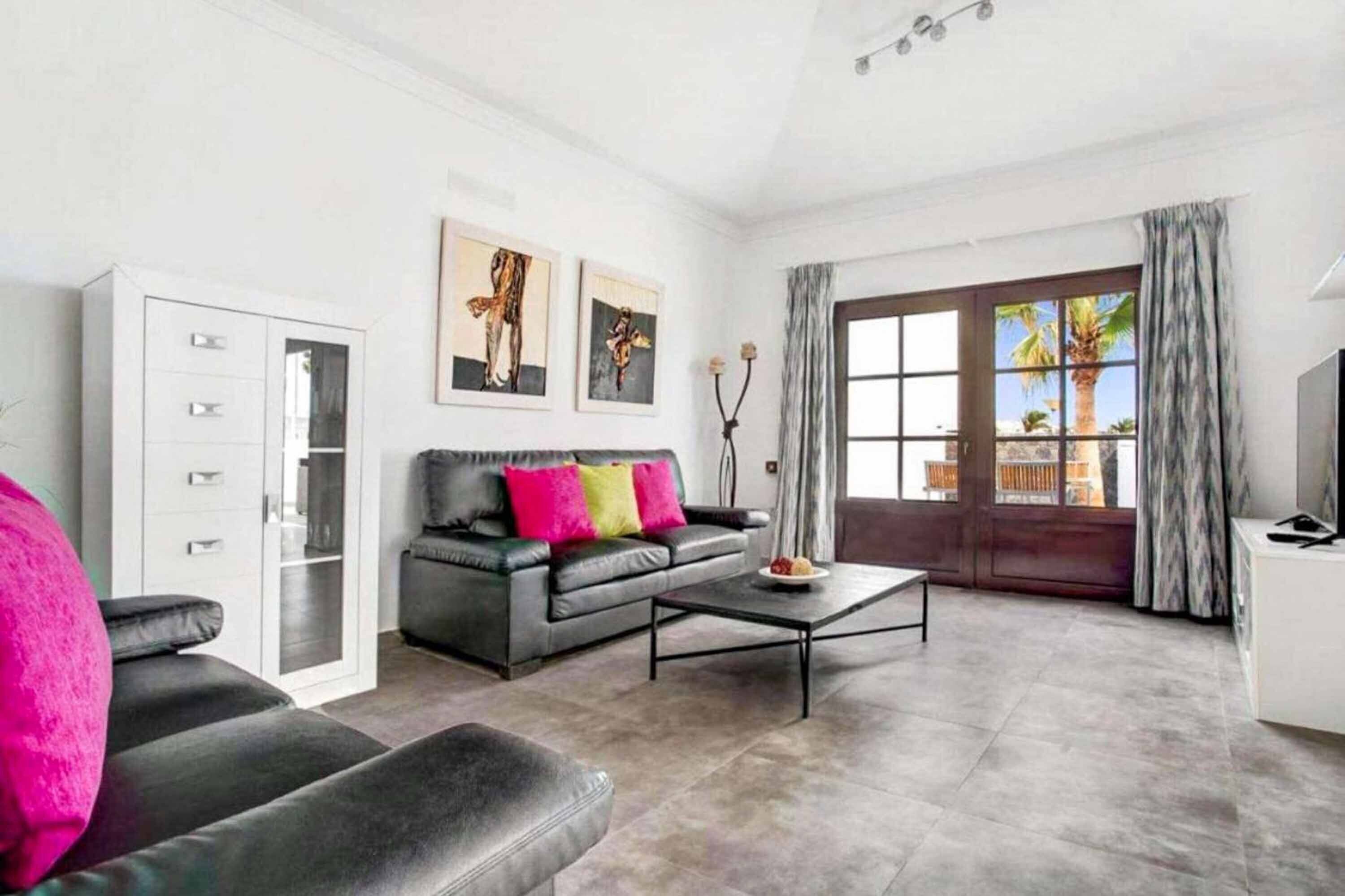 A modern living room with a grey sofa, a coffee table, and a white cabinet. There are two framed artworks on the wall, a large window with curtains, and a door leading outside.
