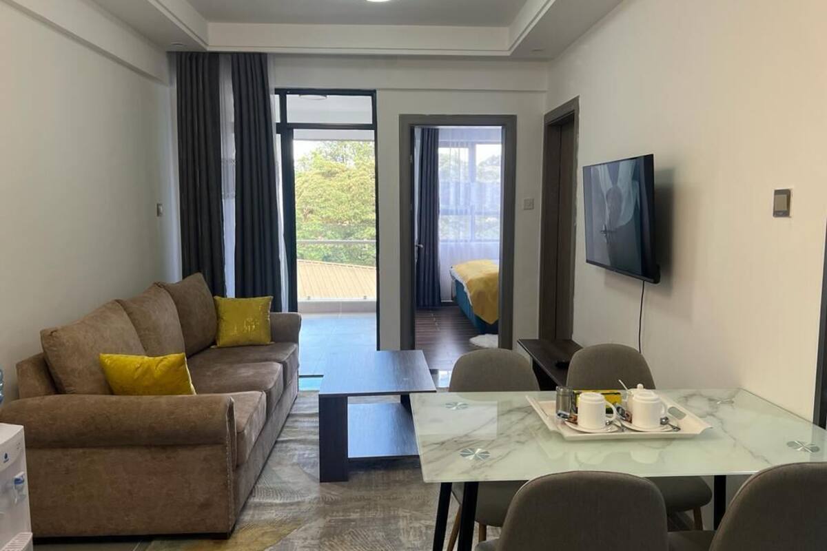 A modern hotel room with a sofa, a dining table with chairs, a flat-screen TV mounted on the wall, and a balcony with a sliding door.