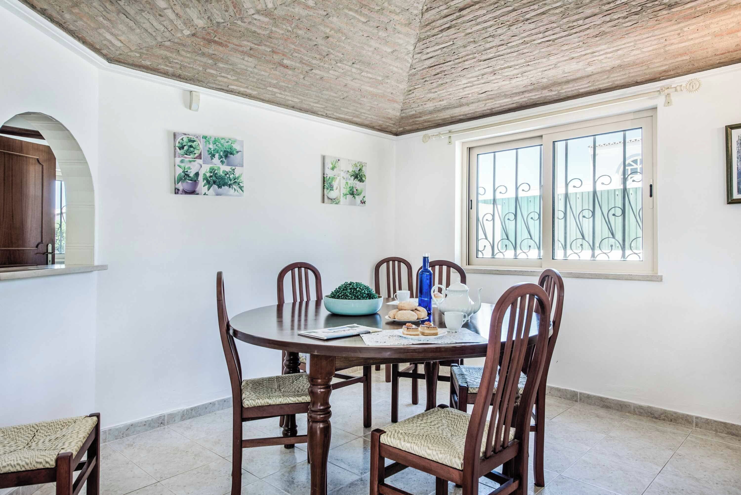 Comedor con una mesa redonda, sillas de madera, un cuenco de fruta, una botella azul y una jarrón blanco.