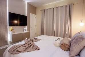 Habitación de hotel con cama, televisor de pantalla plana montado en la pared, mesita de noche con lámpara y ventana con cortinas.