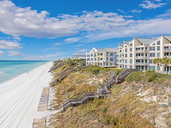 Un hôtel en bord de mer, composé de bâtiments à plusieurs étages, doté d’une plage de sable et d’escaliers en bois menant à la plage.
