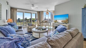 Un salón amplio con un sofá grande, una mesa de centro y un televisor que muestra una escena de playa. Hay un balcón con mobiliario de exterior y vistas al exterior.