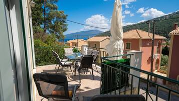 A balcony with outdoor furniture, a table, and chairs, overlooking a scenic view of houses and mountains.