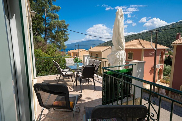 A balcony with outdoor furniture, a table, and chairs, overlooking a scenic view of houses and mountains. - Faos Luxury Apartments 2 (Kefalonia)
