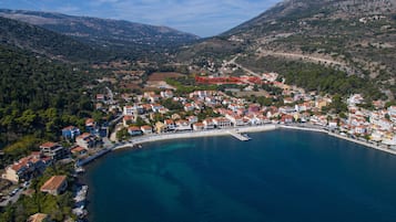 An aerial view of a coastal town with buildings along the shoreline, a clear blue sea, and a mountainous backdrop.