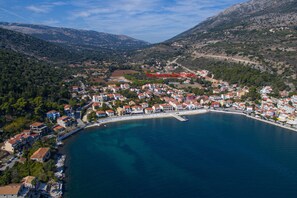 An aerial view of a coastal town with buildings along the shoreline, a clear blue sea, and a mountainous backdrop. - Faos Luxury Apartments 2 (Kefalonia)