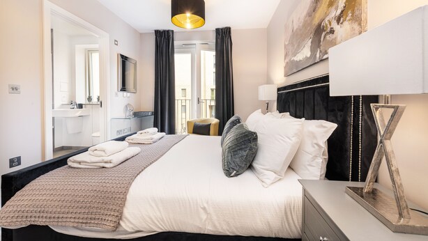 A modern hotel room with a large bed, a bedside table, a mirror, a window with curtains, and a painting on the wall. - Stunning Townhouse XII - Cosy but spacious home from home stays, all in one. (London)