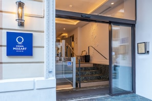 A hotel entrance with a glass door, a blue sign with the hotel name, a staircase, and a wall with a textured pattern. - Crisol Mozart (Nice)