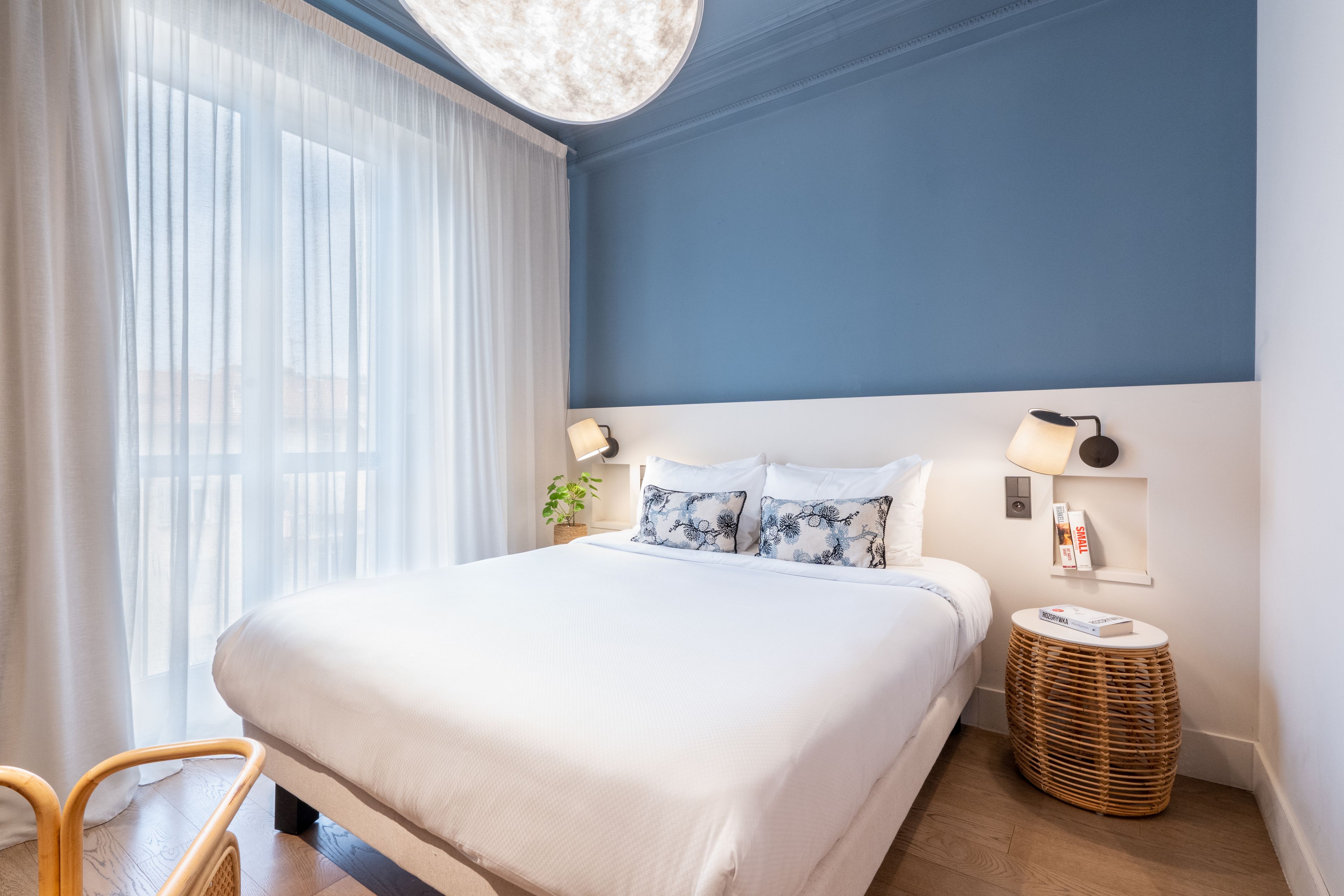 A hotel room with a bed, a bedside lamp, a small table, and a wicker basket.