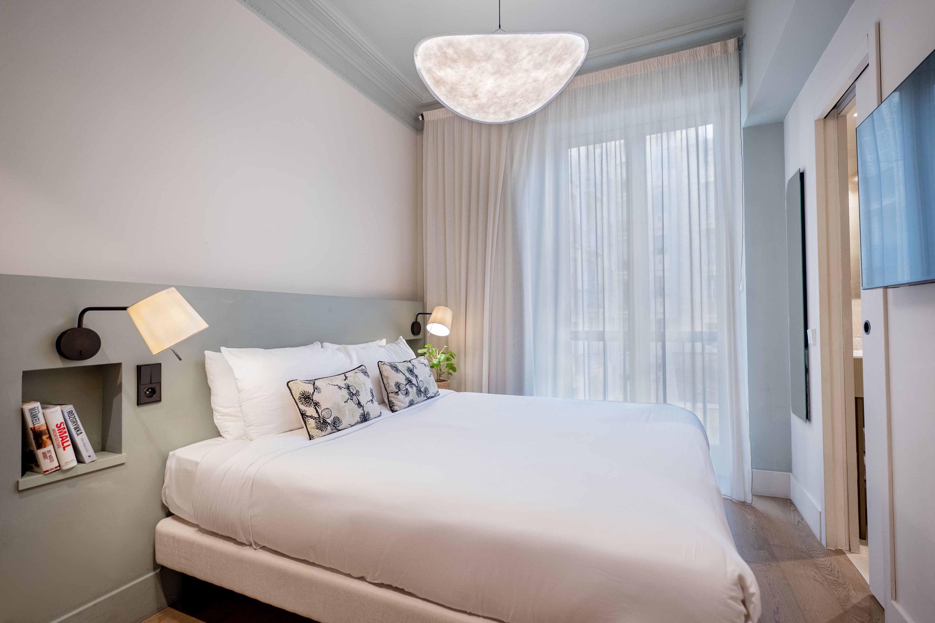 Un dormitorio moderno con una cama grande, una mesita de noche con libros, una lámpara de pared y una ventana con cortinas translúcidas.
