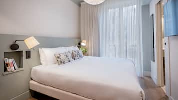 Een moderne slaapkamer met een groot bed, een nachtkastje met boeken, een wandlamp en een raam met dunne gordijnen.