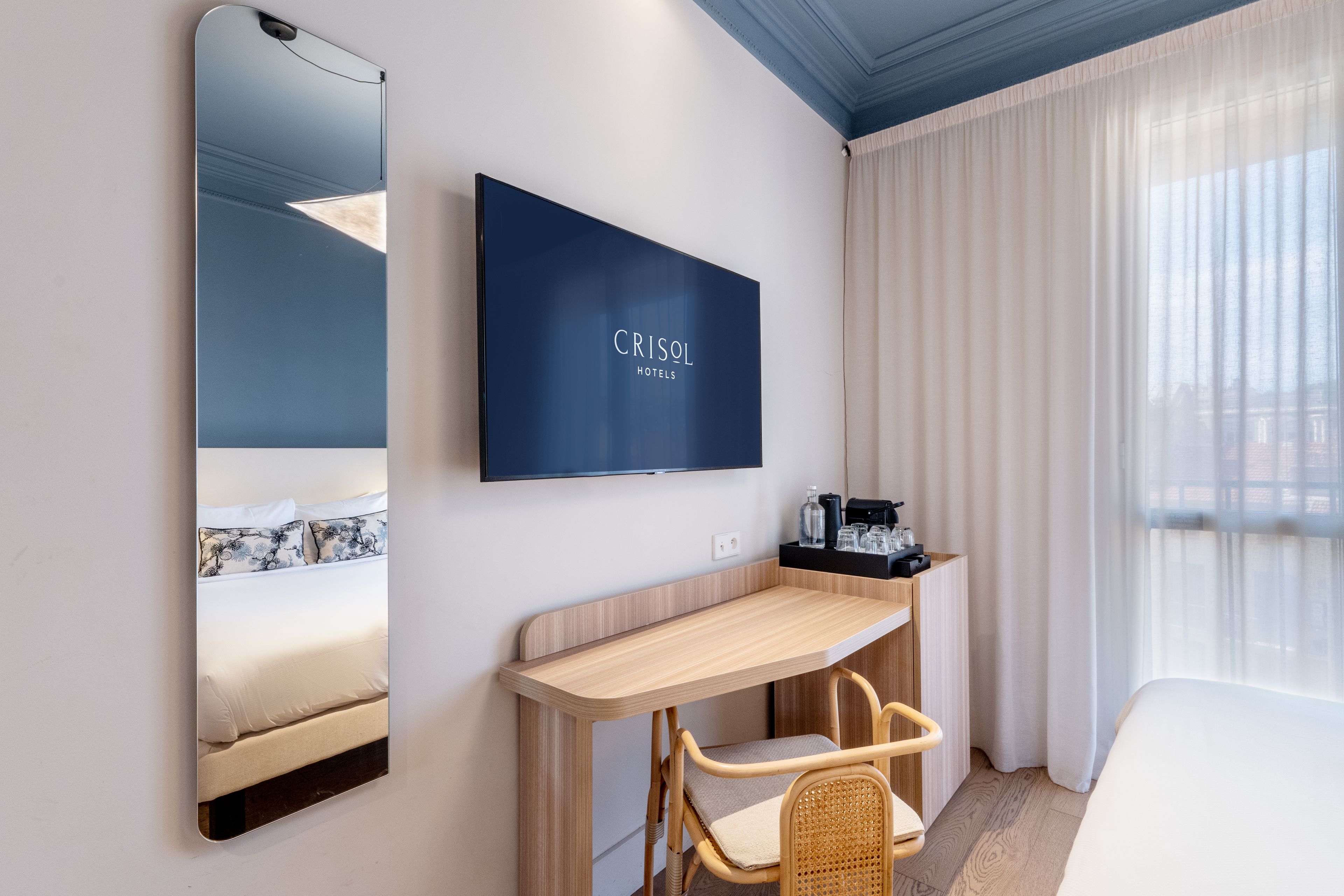 A hotel room with a bed, a TV mounted on the wall, a wooden desk, a wicker chair, and a window with curtains.
