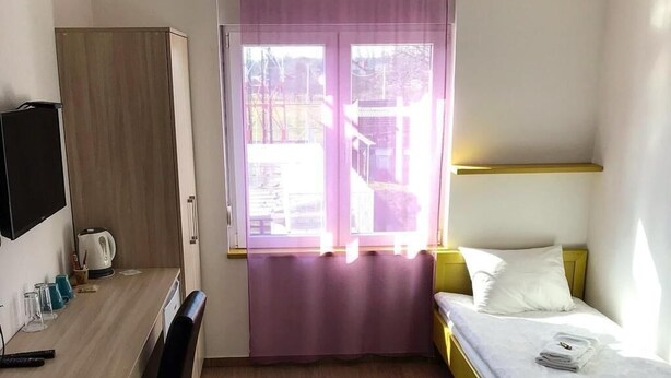 Quarto de hotel com escrivaninha de madeira, cama com colchão branco e janela com cortina roxa.