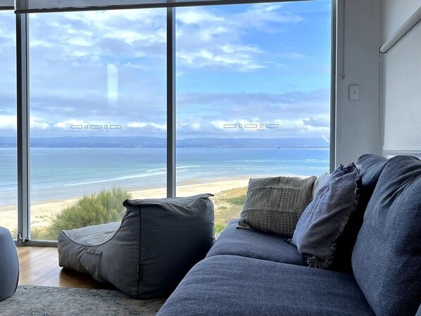 A room with a large window overlooking a beach, a gray sofa with cushions, and a small ottoman.