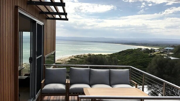 A wooden deck with a sofa and table, overlooking a beach and ocean. - Three Little Birds at Freycinet - Ocean views! (Glamorgan-Spring Bay)