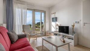Un salon moderne avec un canapé rouge, une table basse blanche, un téléviseur et une vue sur l’extérieur grâce à de grandes portes-fenêtres coulissantes.