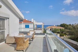 A balcony with wicker furniture, a glass railing, and a view of the sea and neighboring buildings. - Villa Serafi, Stunning 5BDR Kapparis Villa with Private Pool, Close to the Beach (Paralimni)