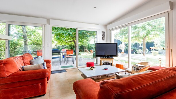 A spacious living room with a red sofa, a coffee table, and a television. Large windows offer a view of the outdoors.