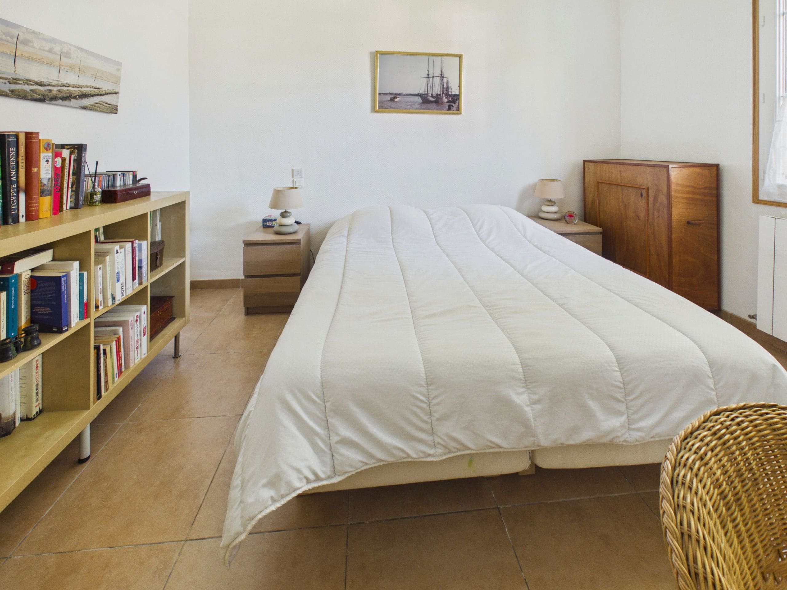 A bedroom with a bed, a wooden bookshelf, a nightstand, a lamp, a framed picture on the wall, and a wicker chair.
