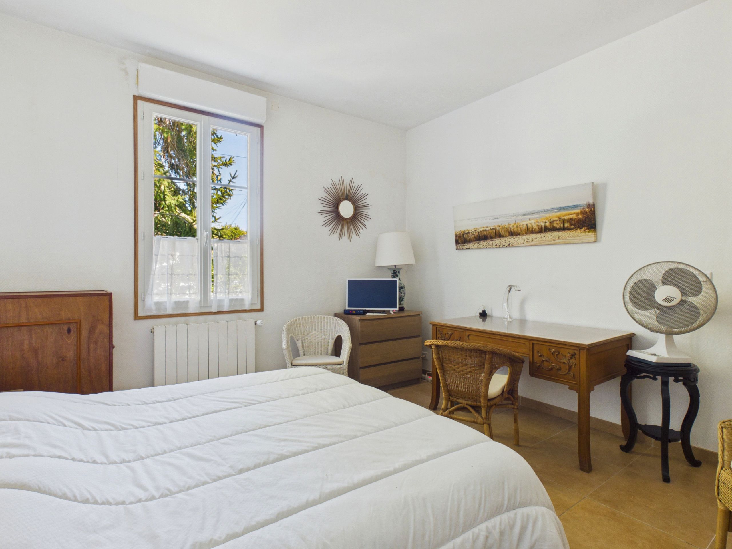 A bedroom with a large bed, a desk, a chair, a computer, a lamp, a fan, and a window with a view of trees.