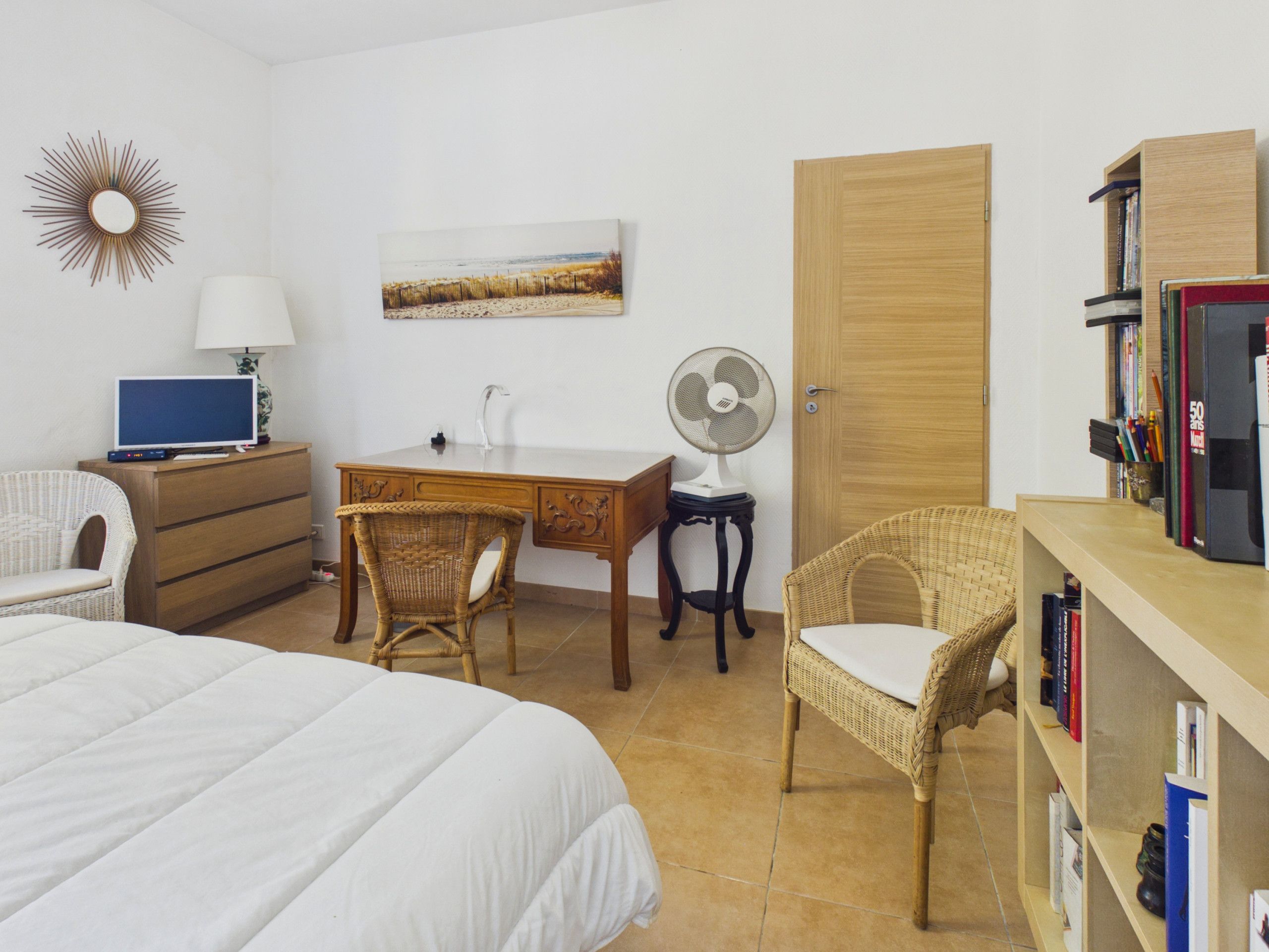 Un dormitorio con cama, escritorio con computadora, silla, ventilador, estantería y una puerta.