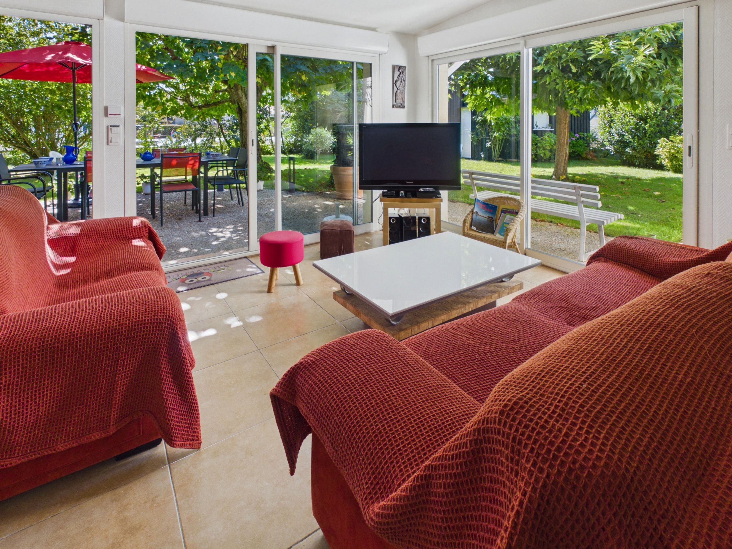 A modern living room with a large TV, red upholstered furniture, and a view of a garden through glass doors.