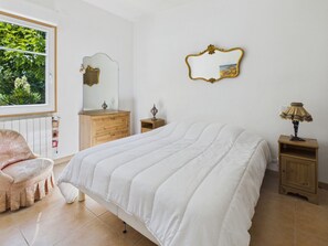 Une chambre à coucher comprenant un lit, une commode, un miroir, une lampe, une chaise et une fenêtre donnant sur un paysage verdoyant.