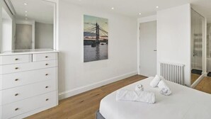 A modern hotel room with a large bed, a dresser with drawers, a wall-mounted mirror, a radiator, and a painting of a bridge.