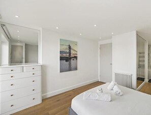 Una habitación de hotel moderna con una cama grande, un aparador con cajones, un espejo fijado a la pared, un radiador y un cuadro de un puente.