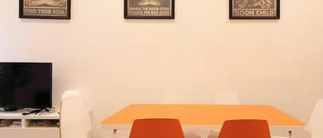 A dining area with a table and chairs, a television on a stand, and three framed posters on the wall.