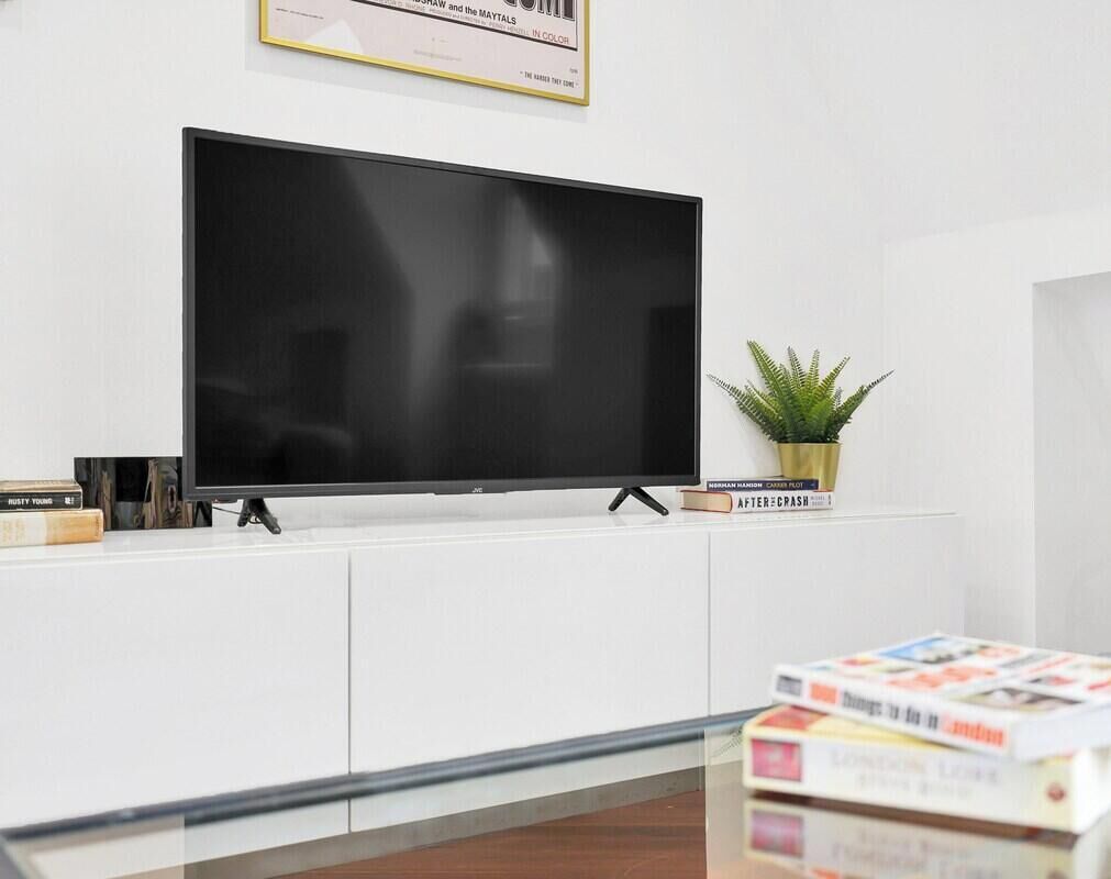 Un salon moderne avec un téléviseur à écran plat fixé à un meuble blanc, une petite plante en pot et une table basse en verre sur laquelle sont posés des livres.