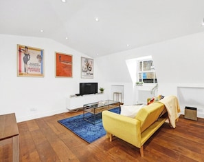 A modern living room with a sofa, coffee table, and a TV. There are posters on the wall and a window with a view.