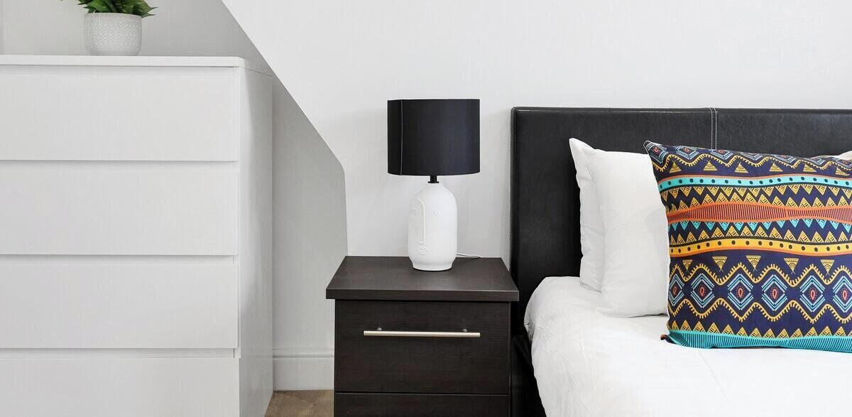 A bedroom with a bed, a nightstand with a lamp, a dresser, and a patterned pillow.