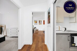 Una habitación de hotel moderna con suelo de madera, paredes blancas, cama, kitchenette y zona de comedor.