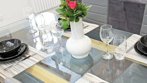 Une table à manger avec un plateau en verre, entourée d’assiettes noires, de verres à vin transparents et d’un vase blanc contenant des fleurs roses.