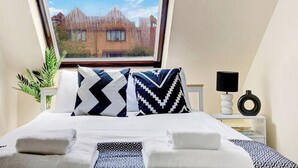 Una cama bien hecha con dos almohadas, una lámpara de noche, una mesita con lámpara y vistas a edificios a través de una ventana.